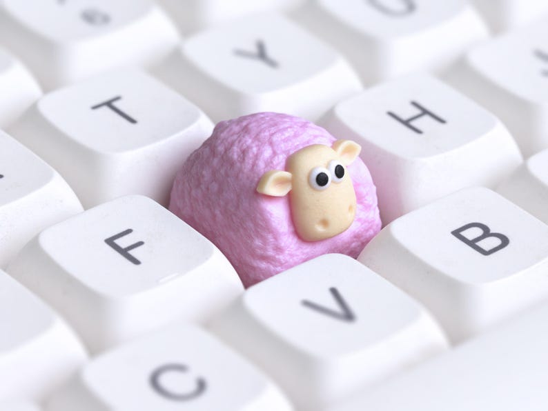 May include: A close-up of a white computer keyboard with a pink sheep-shaped object in the center. The sheep has a light beige face and black eyes. The keyboard keys are labelled with letters.