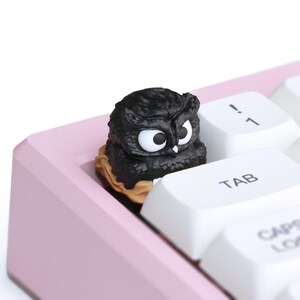 Puede incluir: Una tecla con forma de b&uacute;ho negro se encuentra en un teclado mec&aacute;nico rosa. La tecla tiene grandes ojos blancos y est&aacute; rodeada de teclas blancas con letras negras. El teclado est&aacute; en &aacute;ngulo y el fondo es blanco.