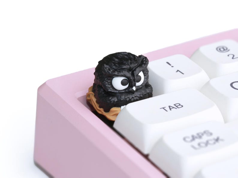 Puede incluir: Una tecla con forma de b&uacute;ho negro se encuentra en un teclado mec&aacute;nico rosa. La tecla tiene grandes ojos blancos y est&aacute; rodeada de teclas blancas con letras negras. El teclado est&aacute; en &aacute;ngulo y el fondo es blanco.