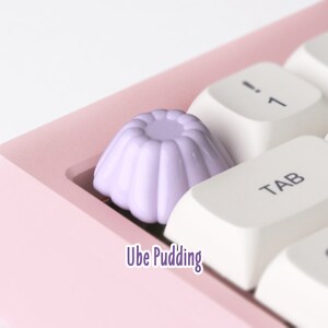 May include: A close-up of a purple keycap shaped like a pudding cup, labeled "Ube Pudding", on a pink keyboard with white keys.
