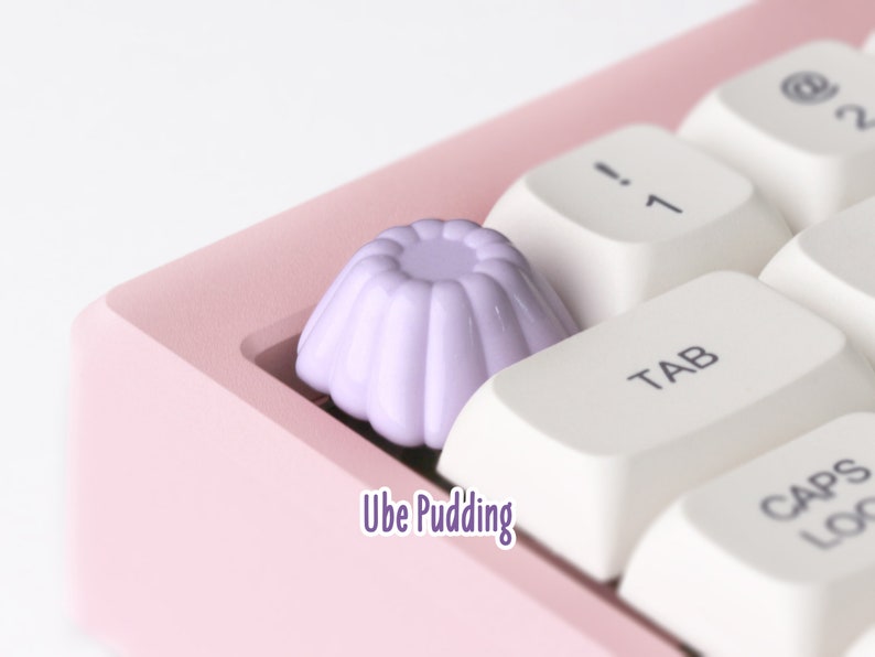 May include: A close-up of a purple keycap shaped like a pudding cup, labeled "Ube Pudding", on a pink keyboard with white keys.