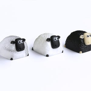 May include: Three sheep figurines, each with a rounded body and a black face. The sheep on the left is white with a textured surface, the middle sheep is white, and the sheep on the right is black.