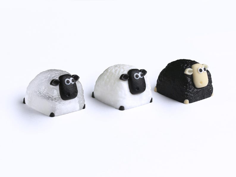 May include: Three sheep figurines, each with a rounded body and a black face. The sheep on the left is white with a textured surface, the middle sheep is white, and the sheep on the right is black.