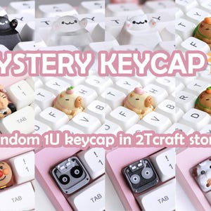 Op de afbeelding: Een roze en wit toetsenbord met een verscheidenheid aan toetsen, waaronder een kat, een hond, een cassettebandje en een cartoonfiguur. De tekst "MYSTERY KEYCAP" wordt weergegeven in roze letters op een witte achtergrond. De tekst "Random 1U keycap in 2Tcraft store)" wordt weergegeven in zwarte letters op een witte achtergrond.