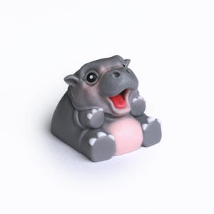 Puede incluir: Una peque&ntilde;a figura de hipop&oacute;tamo gris con la barriga rosa y la boca abierta de par en par, sentada sobre una superficie blanca.