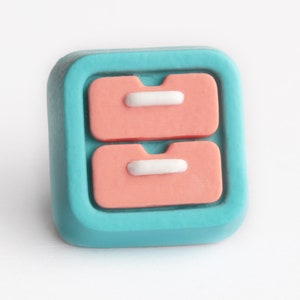 May include: A square-shaped, teal-colored decorative button with a raised design of two stacked, peach-colored drawers. Each drawer has a white, rectangular handle. The button is set against a white background.