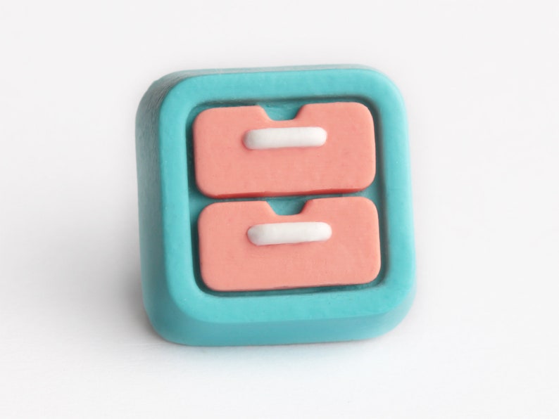 May include: A square-shaped, teal-colored decorative button with a raised design of two stacked, peach-colored drawers. Each drawer has a white, rectangular handle. The button is set against a white background.