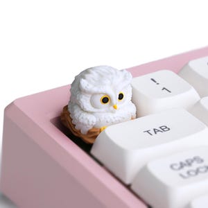 Puede incluir: Una figura de b&uacute;ho blanco con ojos amarillos se encuentra sobre un teclado. El teclado tiene teclas blancas y un marco rosa. El b&uacute;ho est&aacute; posado sobre una base en forma de nido, a&ntilde;adiendo un toque decorativo al teclado.