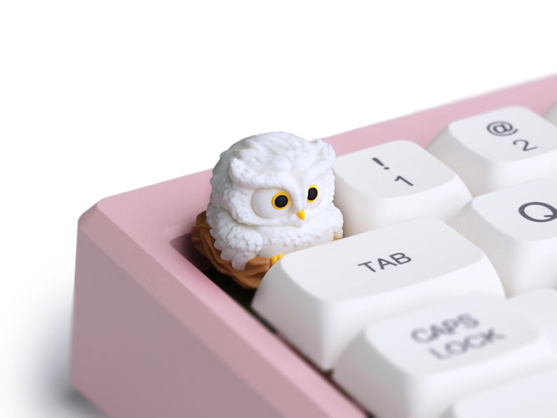Puede incluir: Una figura de b&uacute;ho blanco con ojos amarillos se encuentra sobre un teclado. El teclado tiene teclas blancas y un marco rosa. El b&uacute;ho est&aacute; posado sobre una base en forma de nido, a&ntilde;adiendo un toque decorativo al teclado.