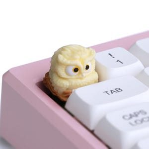 Puede incluir: Una tecla con forma de b&uacute;ho amarillo claro con detalles marrones se encuentra en un teclado rosa. La tecla tiene ojos grandes y redondos y est&aacute; colocada entre teclas blancas. Las teclas del teclado incluyen Tab, Caps Lock y teclas num&eacute;ricas.
