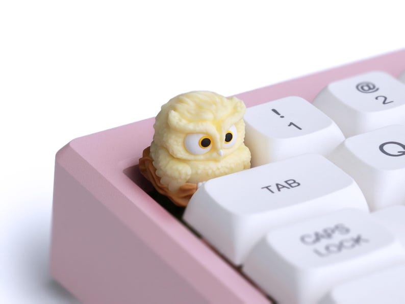 Puede incluir: Una tecla con forma de b&uacute;ho amarillo claro con detalles marrones se encuentra en un teclado rosa. La tecla tiene ojos grandes y redondos y est&aacute; colocada entre teclas blancas. Las teclas del teclado incluyen Tab, Caps Lock y teclas num&eacute;ricas.