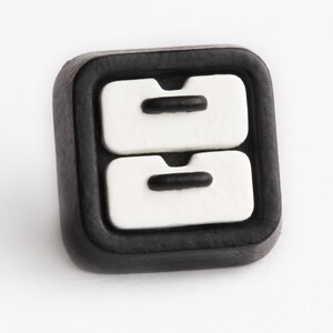 May include: A square, black button with a white design of two stacked drawers. Each drawer has a black handle. The button is likely for clothing or crafts.