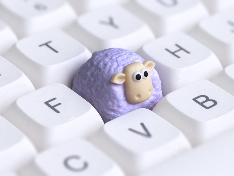 May include: A close-up image of a purple sheep ornament sitting on a white computer keyboard. The sheep has a beige face and ears, with large, cartoonish eyes. The keyboard keys are clean and well-lit, creating a playful contrast.