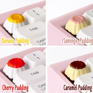 May include: Four different pudding-shaped keycaps for a keyboard. The keycaps are white with a yellow, pink, red, and brown pudding topping. The keycaps are labeled "Banana Pudding", "Flamingo Pudding", "Cherry Pudding", and "Caramel Pudding".
