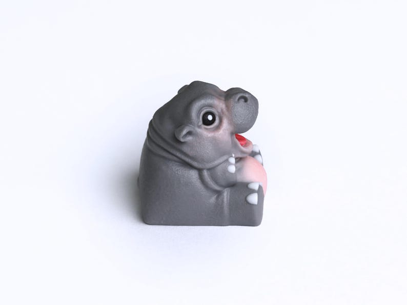 Puede incluir: Una peque&ntilde;a figura de hipop&oacute;tamo gris con la boca rosa y dientes blancos. El hipop&oacute;tamo est&aacute; sentado sobre sus patas traseras con las patas delanteras cruzadas.