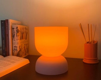 May include: A white and orange ceramic lamp with a round base and a bowl-shaped shade. The lamp is lit and casting a warm orange glow. The lamp is on a wooden table with a book and a decorative vase with sticks.