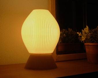 May include: A white, ribbed lampshade with a round base sits on a wooden surface. The lamp is lit and casts a warm glow.