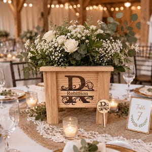 May include: A light-colored wooden planter box featuring a dark engraved monogram with the letter D and the name Robinson. The box is filled with white roses, baby's breath, and eucalyptus greenery. A small matching wooden sign is placed nearby.