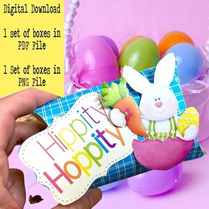 Op de afbeelding: Een blauw-wit geblokte doos met de tekst "Hippity Hoppity" in kleurrijke letters. Een wit konijn dat een wortel vasthoudt, bevindt zich op de doos. De doos is omringd door pastelkleurige plastic paaseieren.