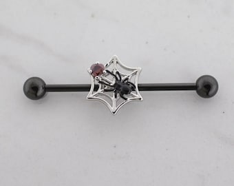 Puede incluir: Una joya corporal de tipo barra industrial negra con un dije de telaraña plateado, una pequeña araña negra y una piedra preciosa redonda de color rojo oscuro. La barra tiene un acabado negro liso con extremos esféricos en ambos lados.