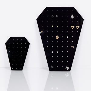 Może przedstawiać: Dwa czarne stojaki na kolczyki z wieloma otworami do przechowywania kolczyków. Większy stojak ma kształt trumny i ma na sobie różne kolczyki, w tym czaszkę, półksiężyc i liść marihuany. Mniejszy stojak jest prostokątny i nie ma na nim żadnych kolczyków.