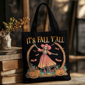 Könnte beinhalten: Schwarze Tragetasche mit dem Text "IT'S FALL Y'ALL". Die Tasche zeigt ein Skelett mit rosa Kleid und Hut, umgeben von Kürbissen und Fledermäusen. Ein dekorativer Seilrahmen umrahmt das Design, perfekt für Halloween.
