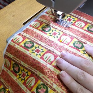 May include: A close-up of a sewing machine needle stitching a red and gold fabric with a Christmas pattern. The fabric has a repeating pattern of candles, holly, and the words "Christmas Holly".