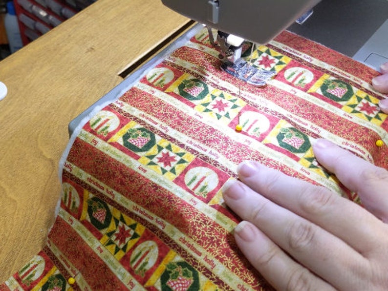 May include: A close-up of a sewing machine needle stitching a red and gold fabric with a Christmas pattern. The fabric has a repeating pattern of candles, holly, and the words "Christmas Holly".