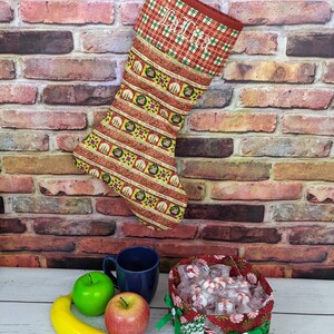 May include: A red and green Christmas stocking with a plaid cuff and the name "Julia" embroidered on it. The stocking is hanging on a brick wall and there is a bowl of sweets and a banana on a white table in the foreground.