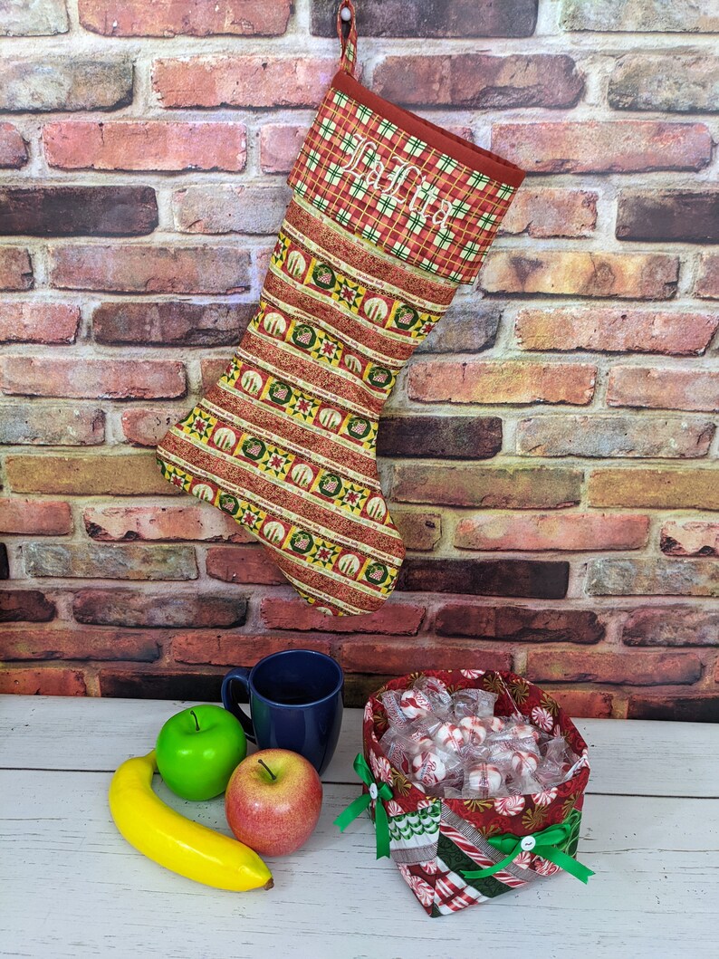 May include: A red and green Christmas stocking with a plaid cuff and the name "Julia" embroidered on it. The stocking is hanging on a brick wall and there is a bowl of sweets and a banana on a white table in the foreground.