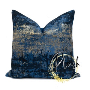 May include: A square decorative pillow with a navy blue and gold abstract design. The pillow has a textured appearance with gold accents. The word "Plush" is written on the pillow in a stylized font.