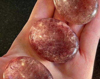 Op de afbeelding: Drie gladde, gepolijste, ovale stenen met een roze en witte wolkachtige uitstraling. De stenen liggen in een hand, de achtergrond is zwart.