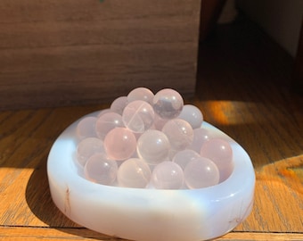 Puede incluir: Un montón de esferas de cuarzo rosa pálido en un plato blanco en forma de corazón. El plato está hecho de piedra pulida.
