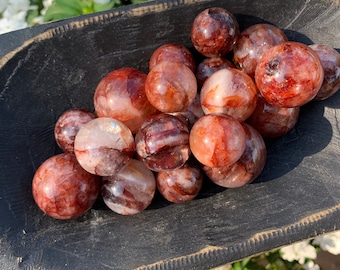 Puede incluir: Una colección de esferas de cristal rojo y blanco, también conocidas como ágata de fuego, en un cuenco de madera. Las esferas son lisas y pulidas, con una variedad de patrones y texturas.