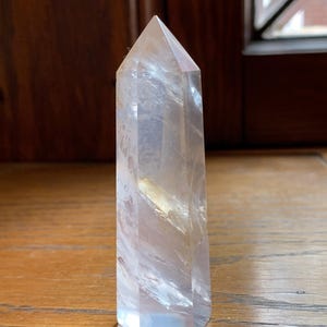 May include: A clear quartz crystal point with a faceted, hexagonal base and a pointed top. The crystal is translucent with internal veining and a slight yellow hue. It stands upright on a wooden surface.