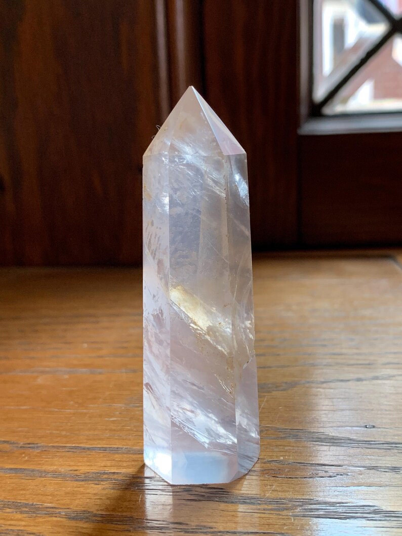 May include: A clear quartz crystal point with a faceted, hexagonal base and a pointed top. The crystal is translucent with internal veining and a slight yellow hue. It stands upright on a wooden surface.