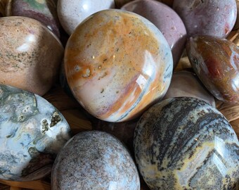 Può includere: Una collezione di pietre di agata multicolori lucidate in un cesto intrecciato. Le pietre sono rotonde e presentano una varietà di motivi e colori, tra cui marrone, bianco, grigio e rosso.