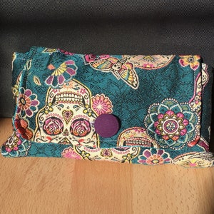 Peut inclure: Une pochette en tissu bleu sarcelle avec un motif color&eacute; de cr&acirc;ne de sucre du Jour des Morts. La pochette a une fermeture &agrave; bouton violet et est d&eacute;cor&eacute;e de fleurs roses, jaunes et blanches.
