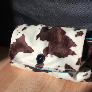 Peut inclure: Une pochette en tissu &agrave; imprim&eacute; vache blanc et marron avec une fermeture &agrave; bouton noir. La pochette est bord&eacute;e de turquoise.