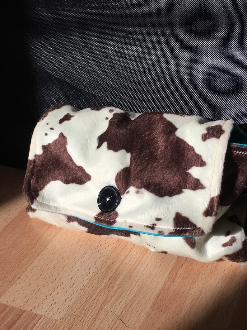 Peut inclure: Une pochette en tissu &agrave; imprim&eacute; vache blanc et marron avec une fermeture &agrave; bouton noir. La pochette est bord&eacute;e de turquoise.