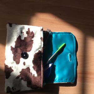 Peut inclure: Une pochette de soir&eacute;e en tissu imprim&eacute; vache, marron et blanc, avec un bouton noir. La pochette est ouverte et l'int&eacute;rieur est doubl&eacute; d'un tissu turquoise brillant. Un stylo vert et un surligneur bleu se trouvent &agrave; l'int&eacute;rieur de la pochette.