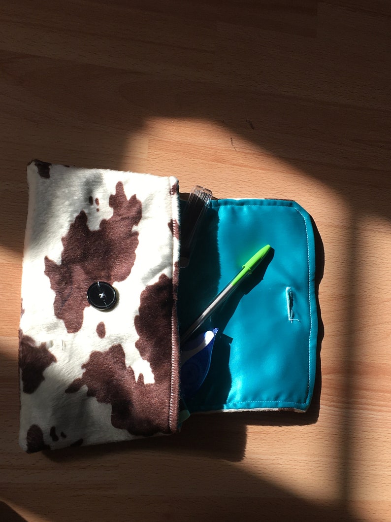 Peut inclure: Une pochette de soir&eacute;e en tissu imprim&eacute; vache, marron et blanc, avec un bouton noir. La pochette est ouverte et l'int&eacute;rieur est doubl&eacute; d'un tissu turquoise brillant. Un stylo vert et un surligneur bleu se trouvent &agrave; l'int&eacute;rieur de la pochette.