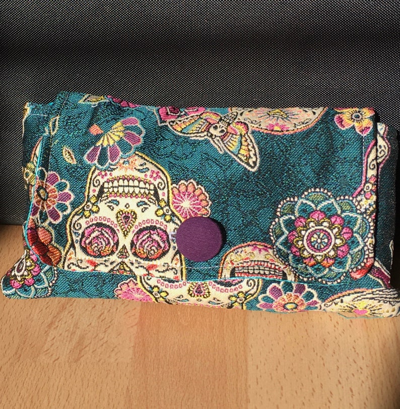 Peut inclure: Une pochette en tissu bleu sarcelle avec un motif color&eacute; de cr&acirc;ne de sucre du Jour des Morts. La pochette a une fermeture &agrave; bouton violet et est d&eacute;cor&eacute;e de fleurs roses, jaunes et blanches.