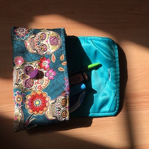 Peut inclure: Une pochette en tissu bleu sarcelle avec une fermeture &agrave; glissi&egrave;re et un motif floral et cr&acirc;ne color&eacute;. La pochette est ouverte et a une doublure bleu turquoise. La pochette est remplie d'un surligneur vert, d'un stylo bleu et d'une gomme blanche et bleue.