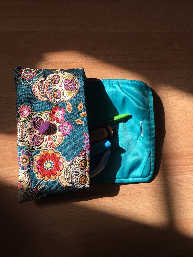 Peut inclure: Une pochette en tissu bleu sarcelle avec une fermeture &agrave; glissi&egrave;re et un motif floral et cr&acirc;ne color&eacute;. La pochette est ouverte et a une doublure bleu turquoise. La pochette est remplie d'un surligneur vert, d'un stylo bleu et d'une gomme blanche et bleue.