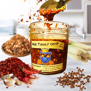 May include: A glass jar filled with a red and orange chili oil with a label that reads "PKR TINGLY CRISP" and lists the ingredients: Sichuan peppercorn, Thai chili, red chili flake, garlic, lemongrass, onion, salt, sugar, oil.