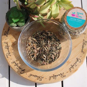 May include: A clear glass bowl filled with dried herbs and spices sits on a wooden cutting board. A small potted succulent and a jar with a cork lid are also on the board. The jar has a label that reads "Down to Earth Zenergy Pased". The board is engraved with the words "Blessed, Grateful, Thankful".