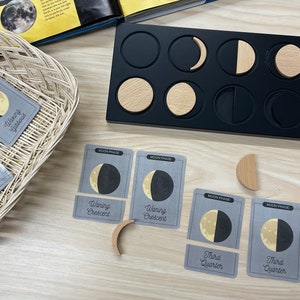Puede incluir: Una bandeja de madera negra con recortes circulares contiene formas de fases lunares de madera. Tarjetas grises con ilustraciones y texto de fases lunares están dispersas, junto con una cesta tejida.