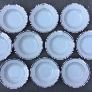May include: Set of ten white ceramic soup bowls with blue and gold trim. The bowls are arranged in a circle on a grey surface.