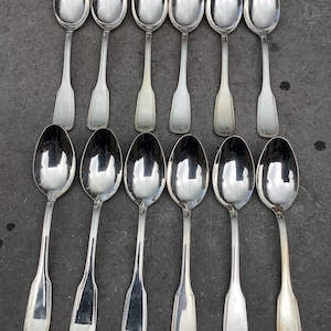 Puede incluir: Un juego de doce cucharas de plata con mangos ornamentados. Las cucharas están dispuestas en dos filas de seis, con los mangos apuntando hacia el centro de la imagen.